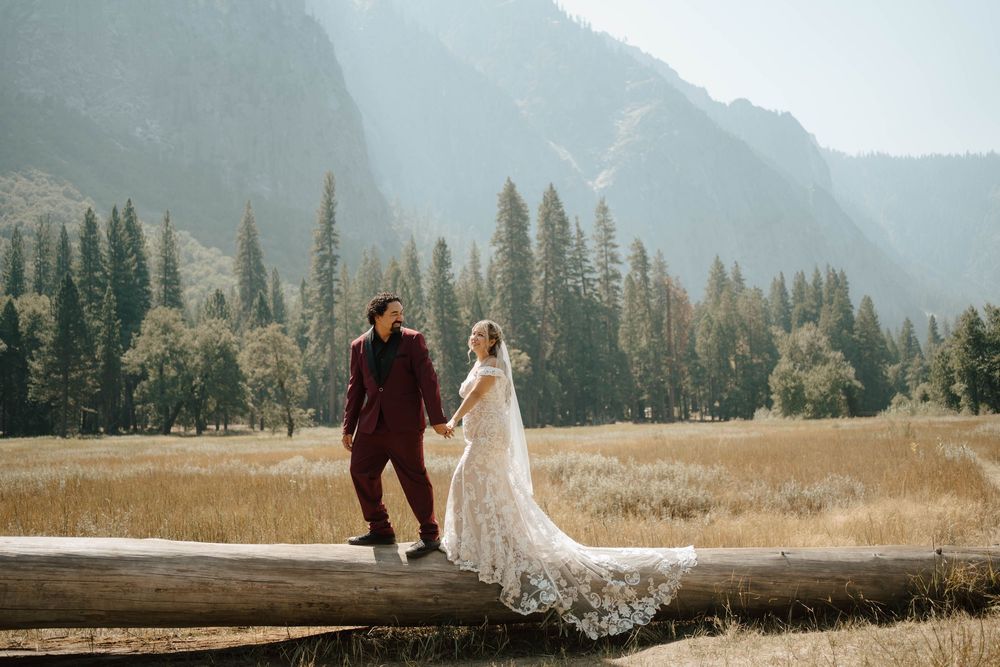 Bride and groom holding hands while balancing on a fallen log in a misty Yosemite meadow.