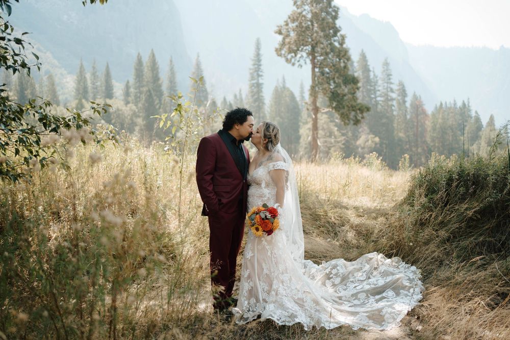 Couple kissing in golden meadow with bride holding colorful fall bouquet and dramatic mountain backdrop.
