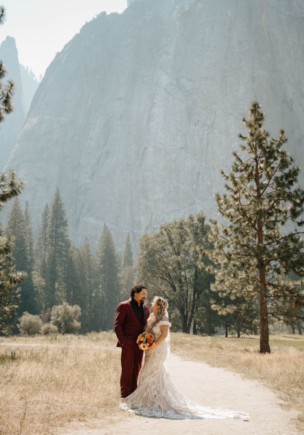 Bride and groom standing close together on a path with towering Yosemite granite cliffs behind them.
