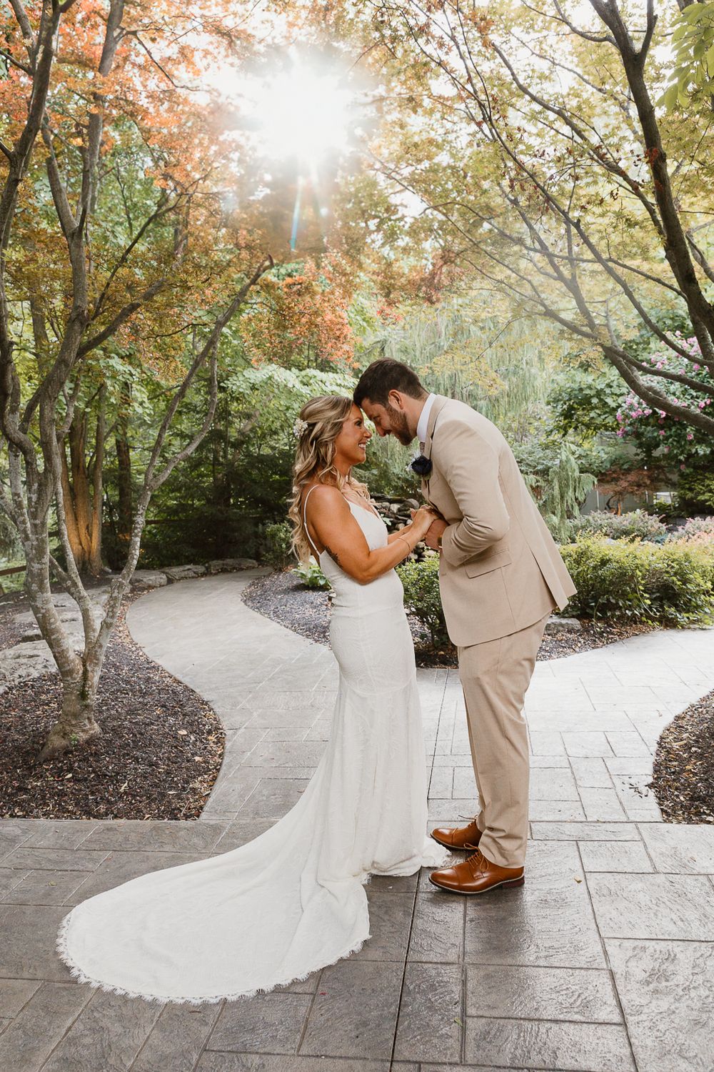 A romantic wedding couple embraces on a stone path surrounded by autumn trees with sunlight streaming through branches.