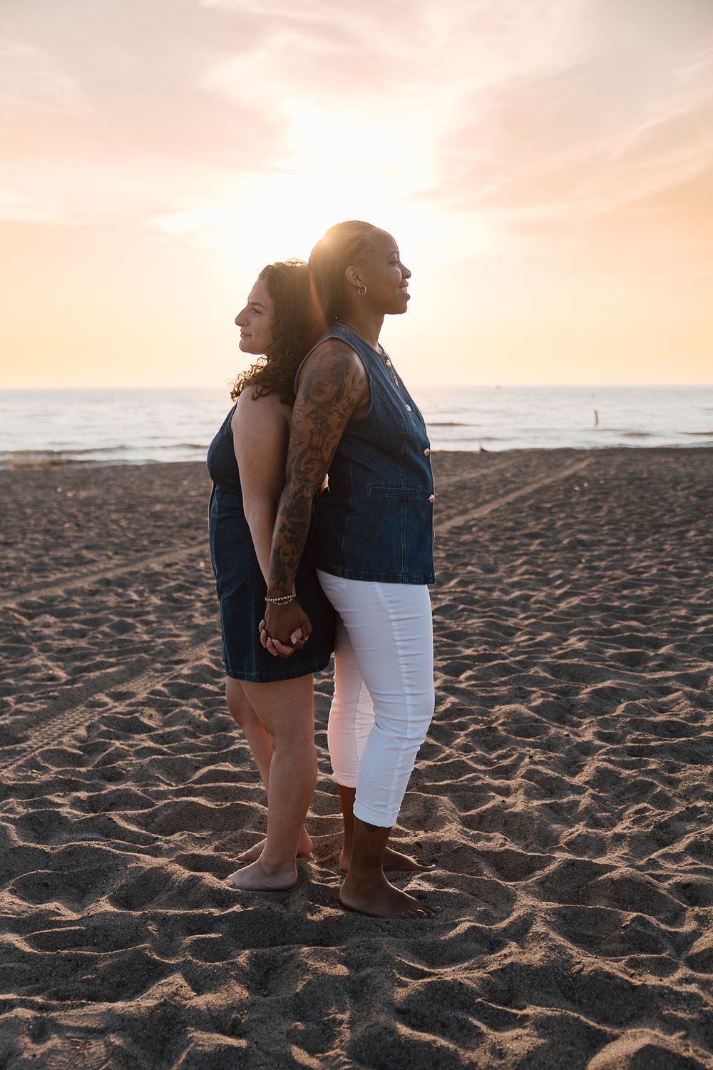 A couple stands back-to-back on a sandy beach at sunset, holding hands while looking out at the ocean.