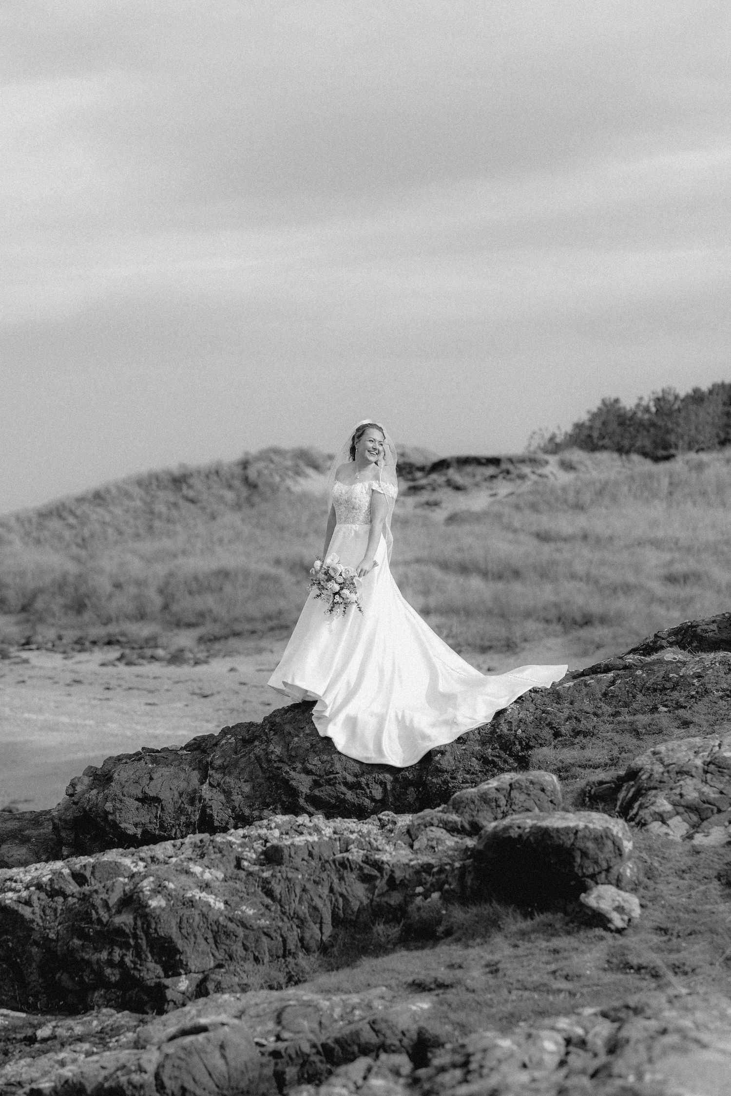 A flowing white wedding dress billows dramatically against rocky coastal cliffs in black and white photography.