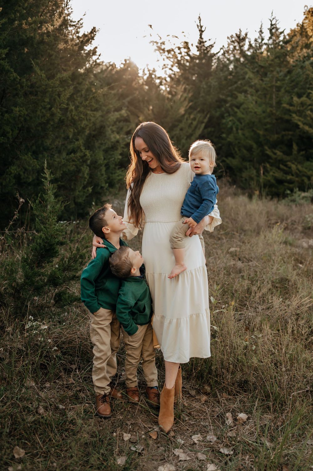 Mother holding baby while young son gives a kiss in a golden hour outdoor setting.