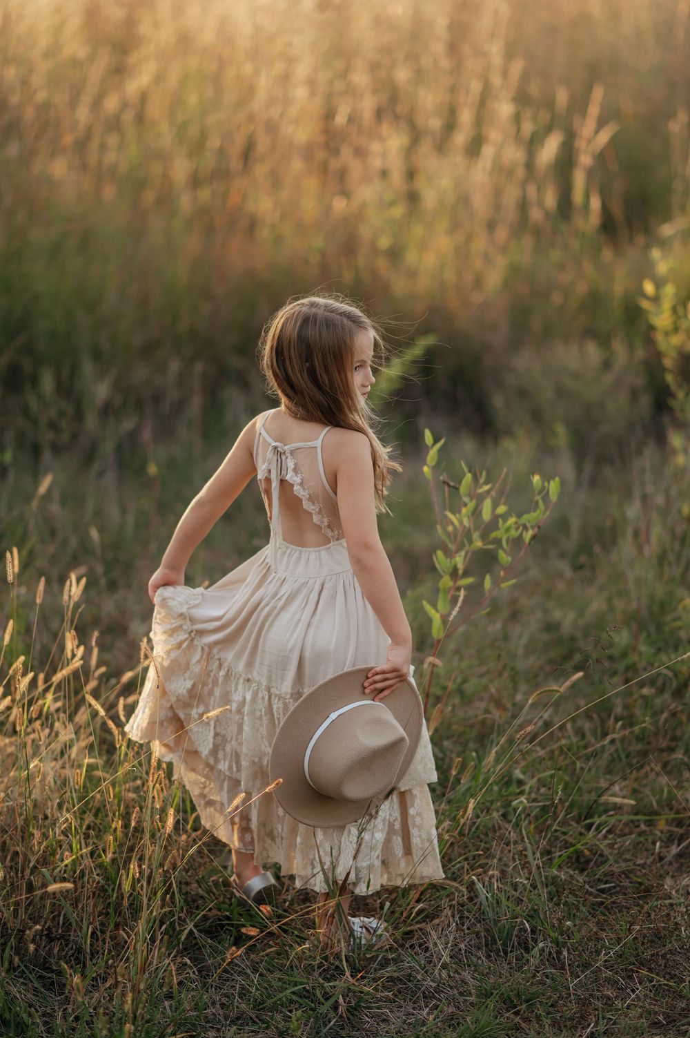 Young girl in a boho lace dress holding a hat while walking through a sunlit field.
