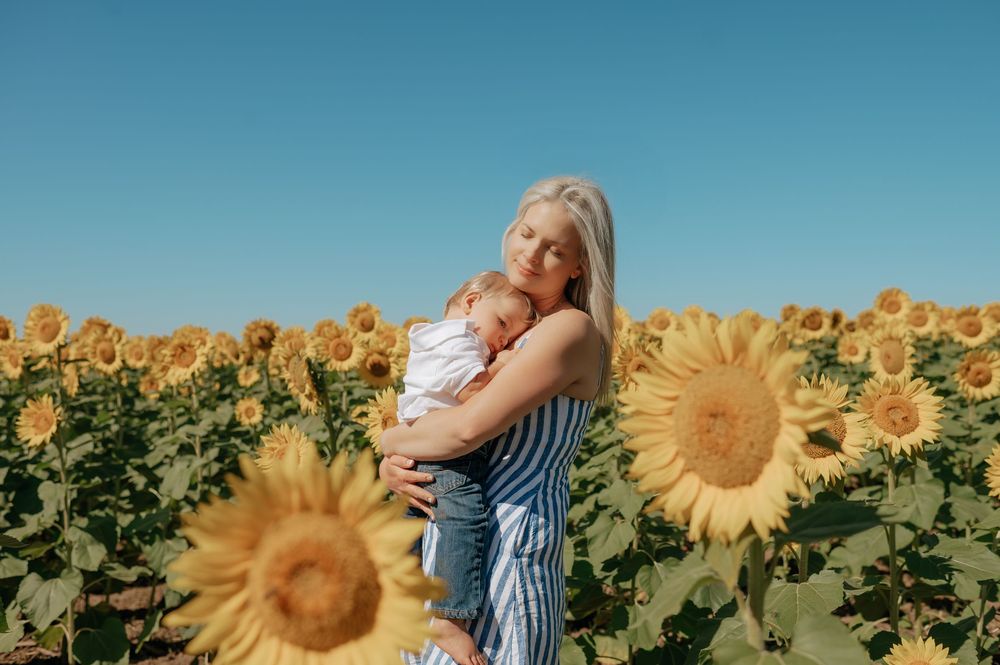 Woman with white hair holding baby in sunflower field under blue sky.