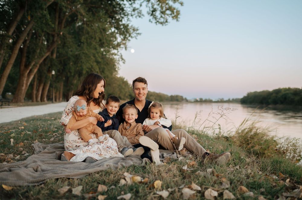 A happy family of six relaxes together on a blanket beside a calm river at dusk.