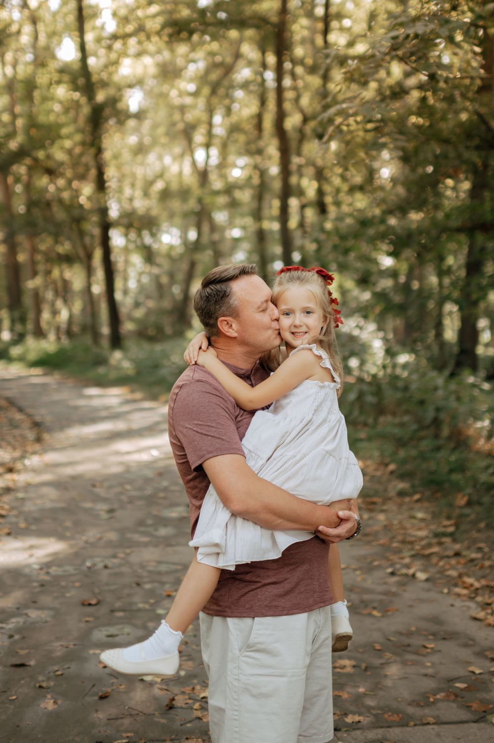 Father kisses his young daughter on the cheek while holding her in a sunny forest path.