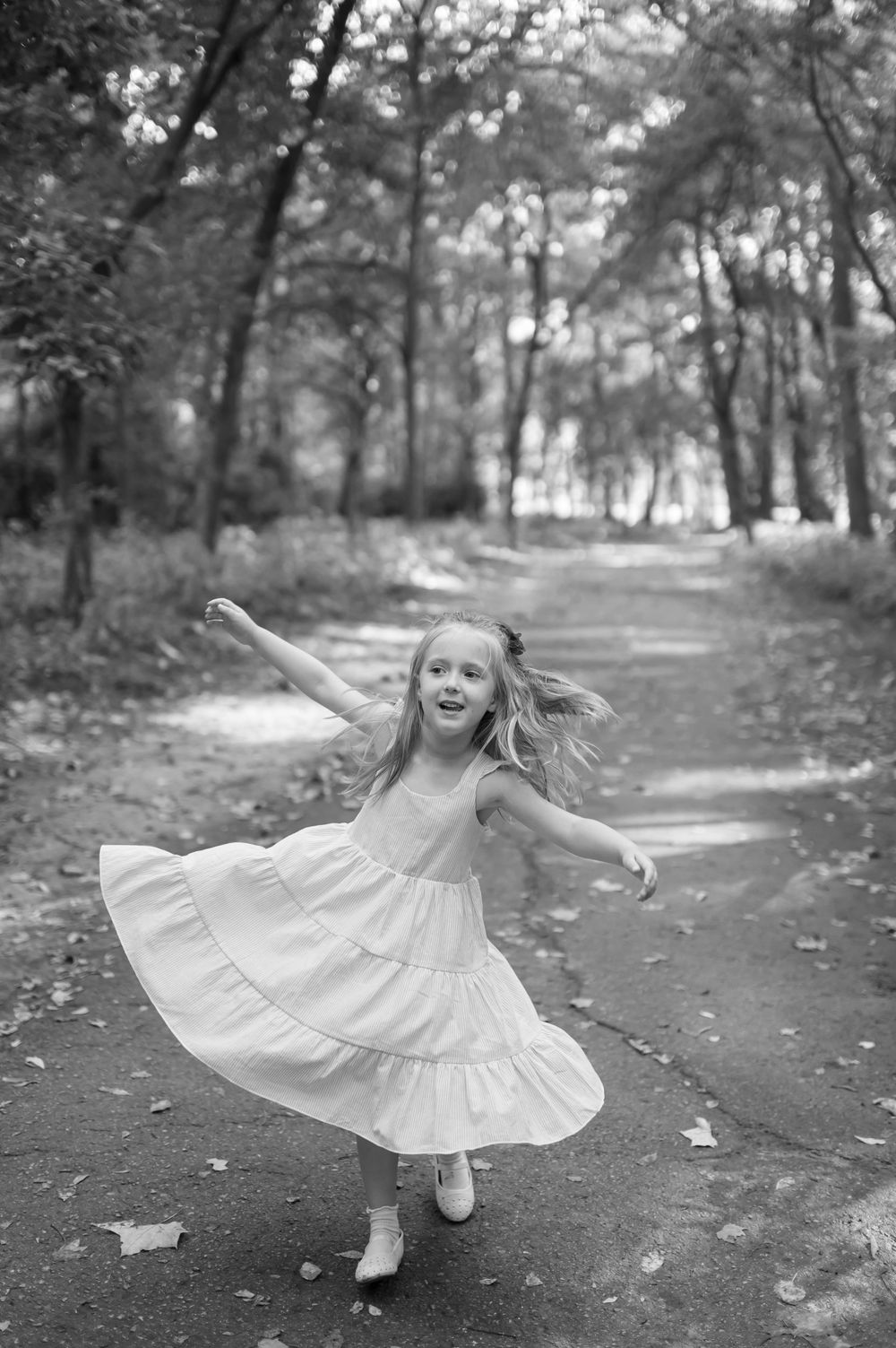 A joyful young girl in a white dress twirls on a tree-lined path in a black and white outdoor portrait.