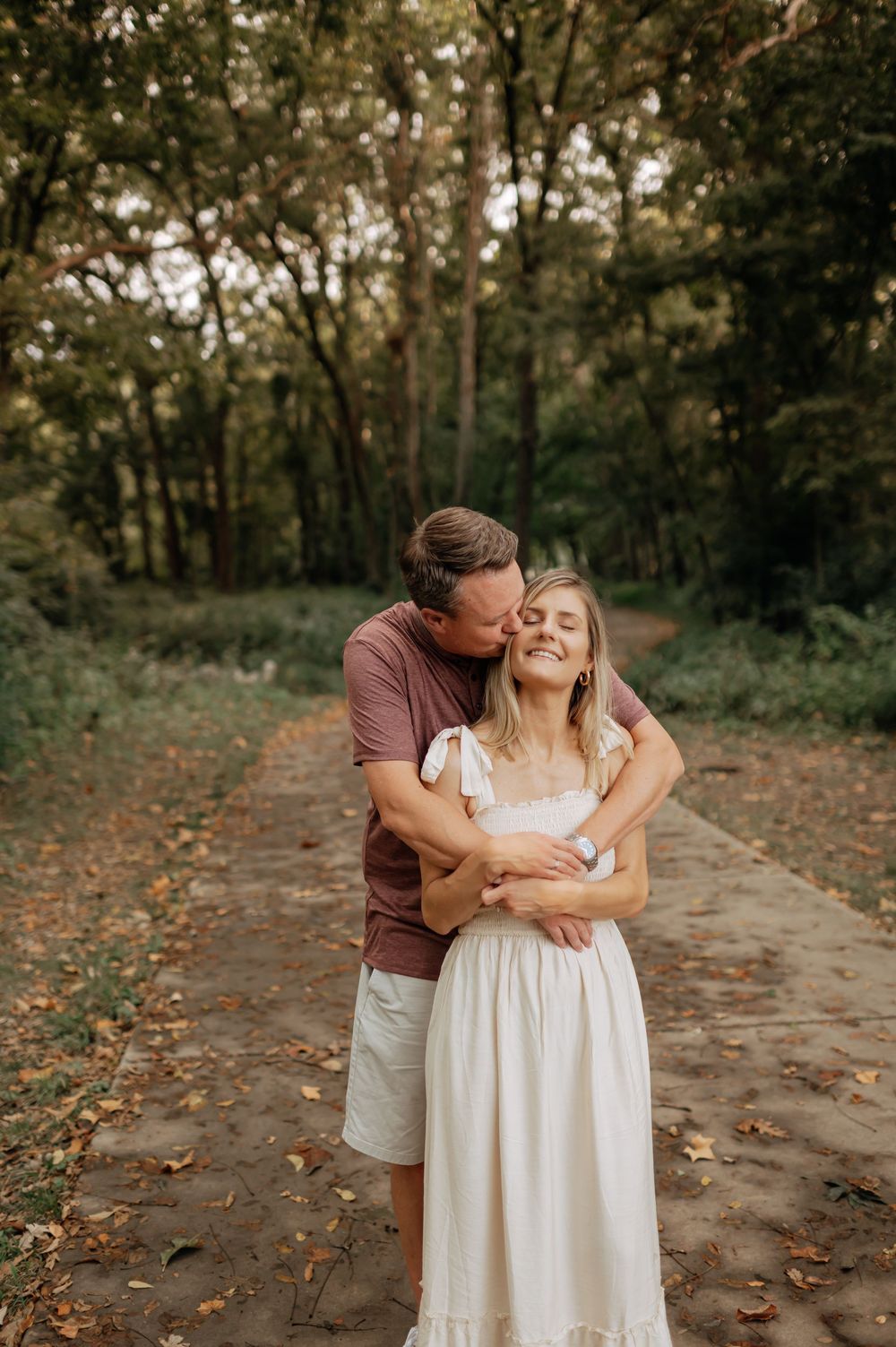 A couple shares a tender embrace on a forest path, the man kissing the smiling woman's cheek.