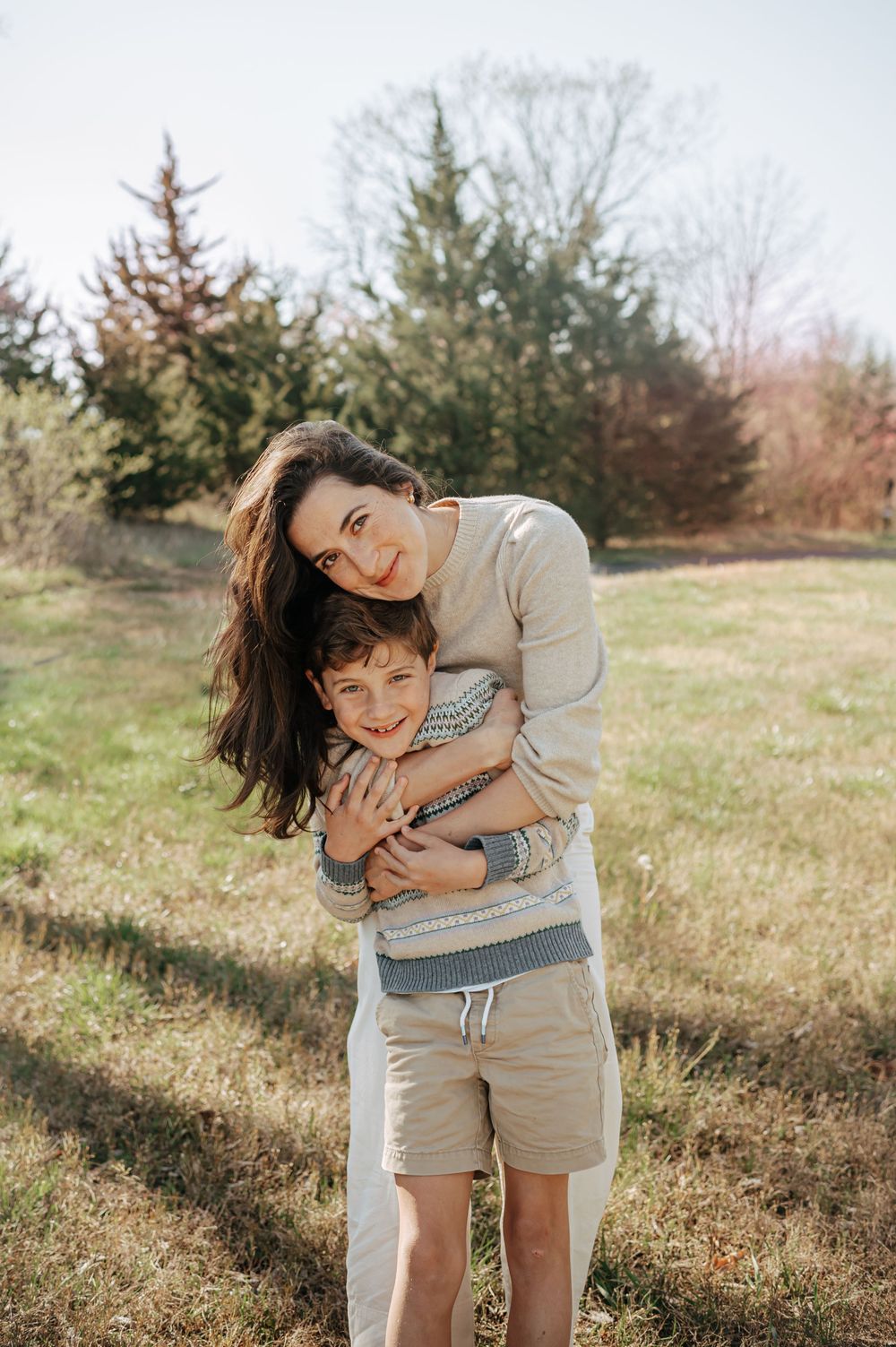 A dark-haired mother hugs her smiling young son from behind in a sunny open field.