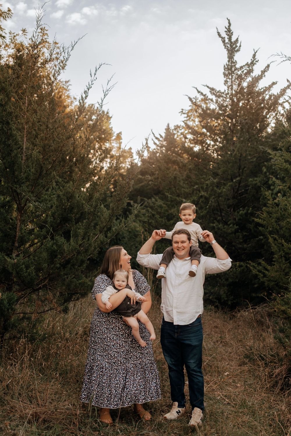 A young family poses outdoors with dad carrying toddler on shoulders and mom holding a baby.