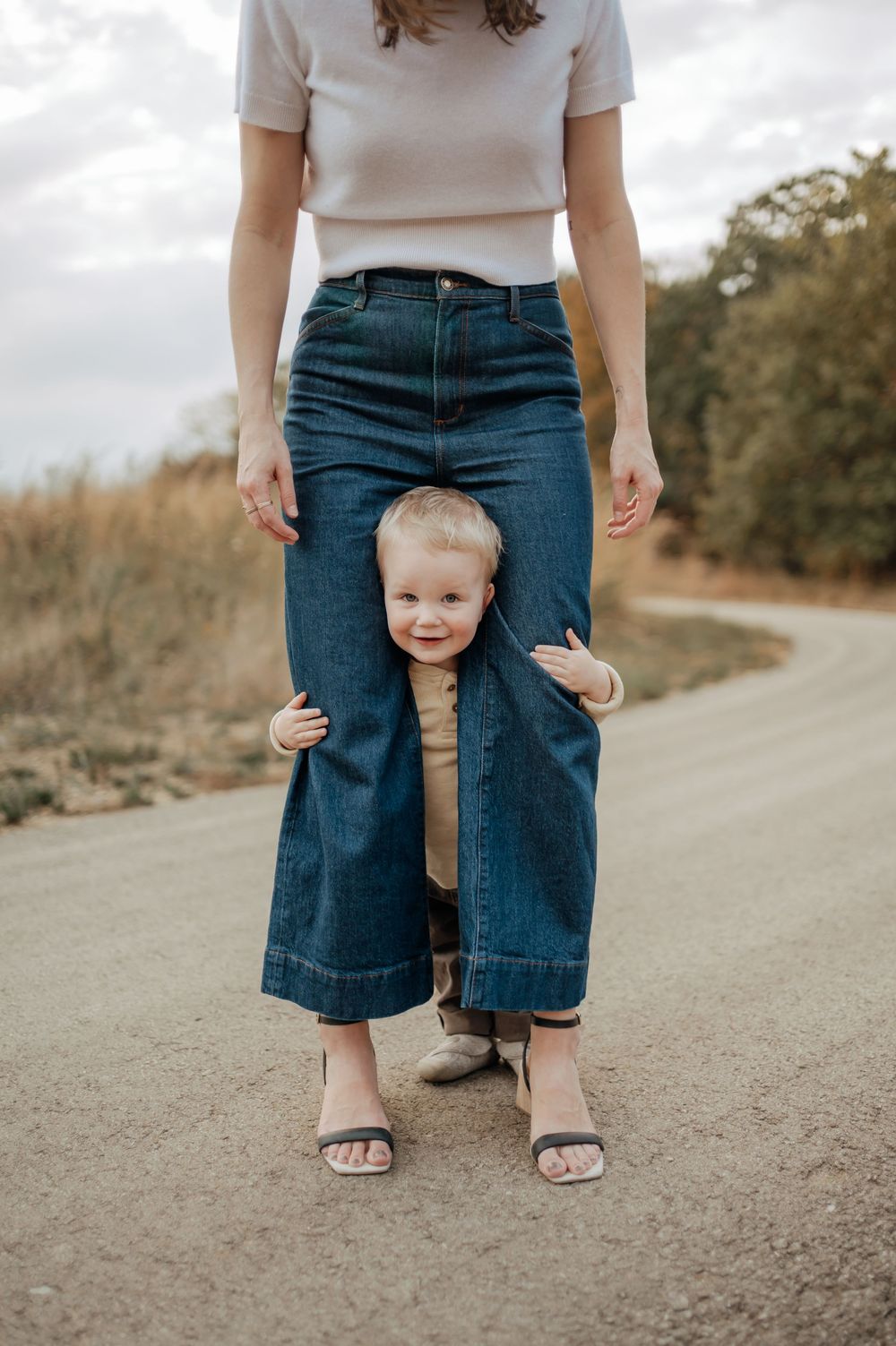 Curious toddler peeking through mother's wide-leg jeans while standing on a rural dirt road.