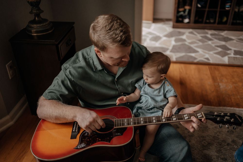 Father playing acoustic guitar while holding infant on lap, sitting on hardwood floor at home.