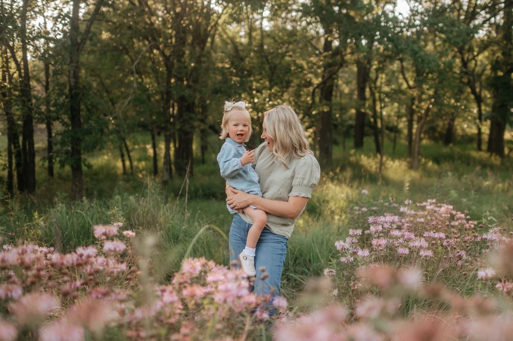 Blonde mother holding her toddler daughter among pink wildflowers in a lush green forest.