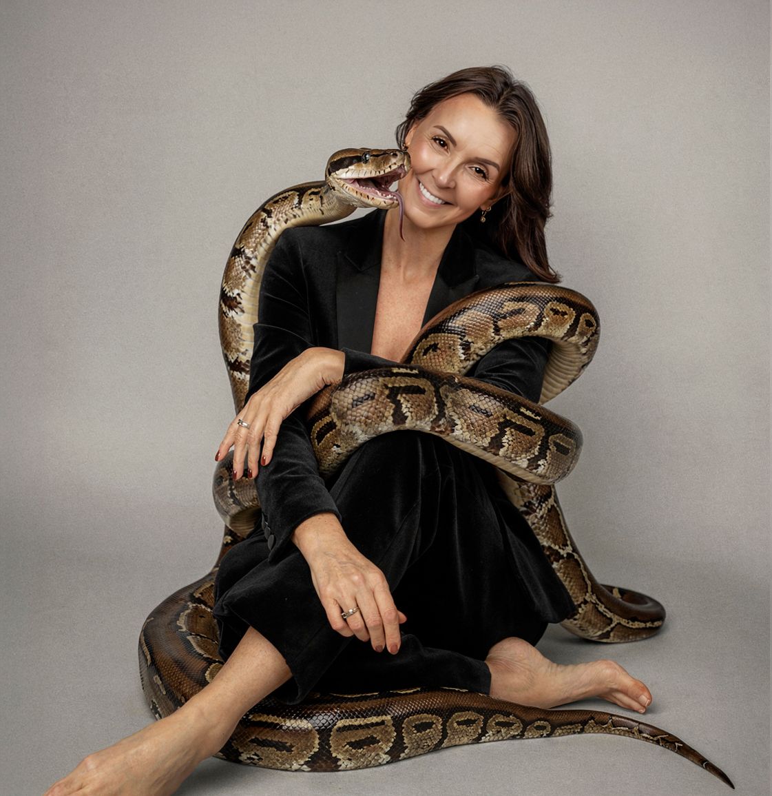 A photoshoot featuring someone in black clothing posing with a large brown and tan python snake against a gray backdrop.