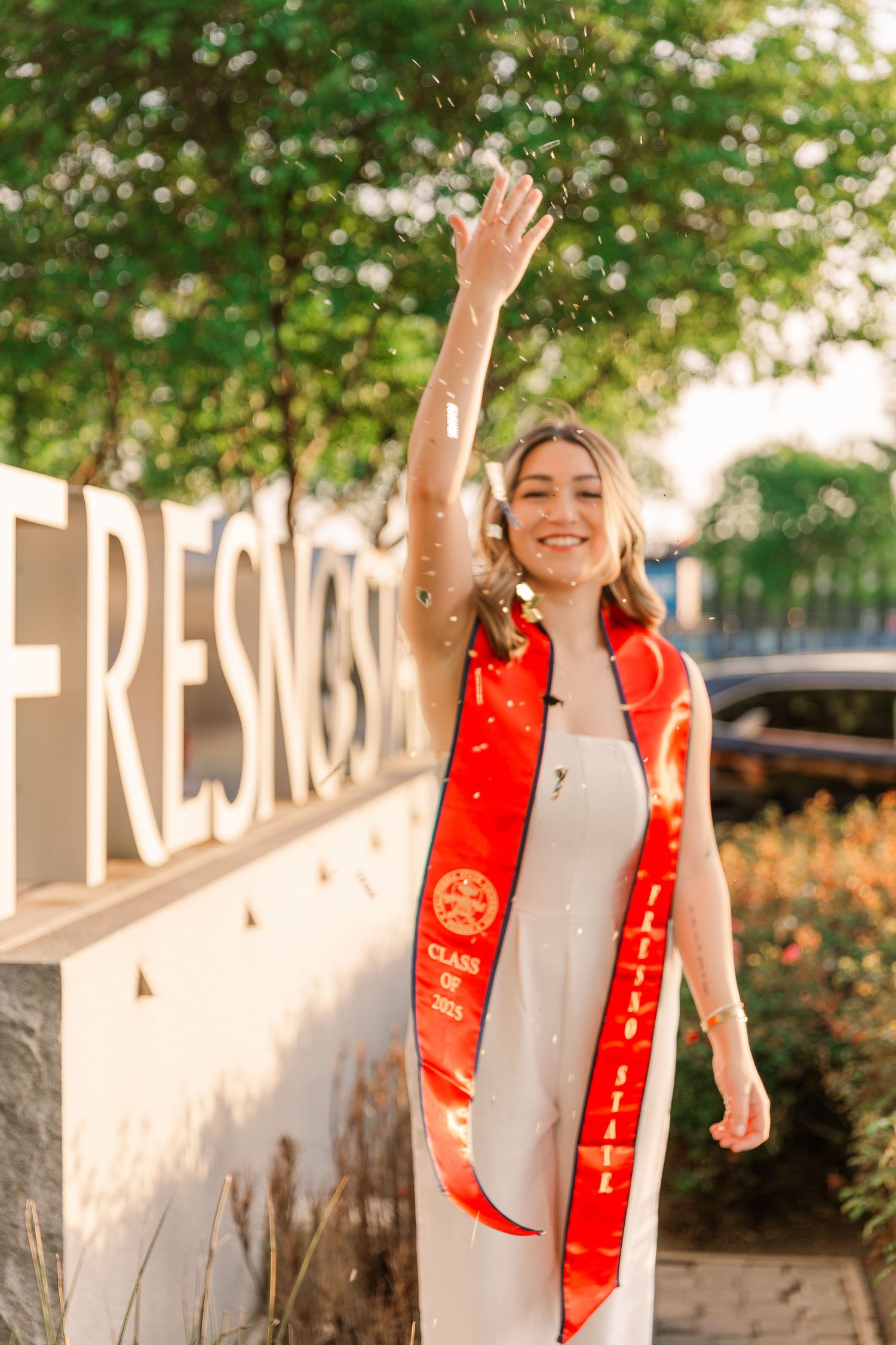 A person in a white dress and red sash poses outdoors against a white wall with greenery in the background.