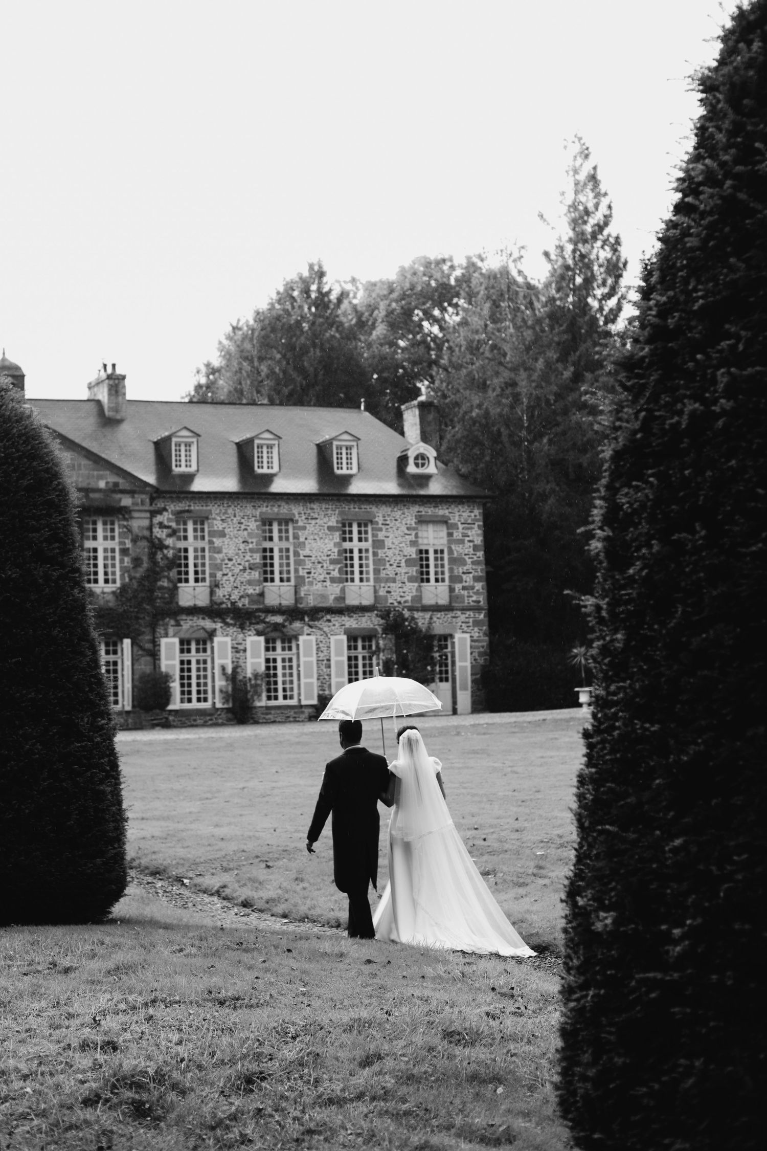 Black and white photo of bride and groom walking with umbrella toward elegant stone manor house.