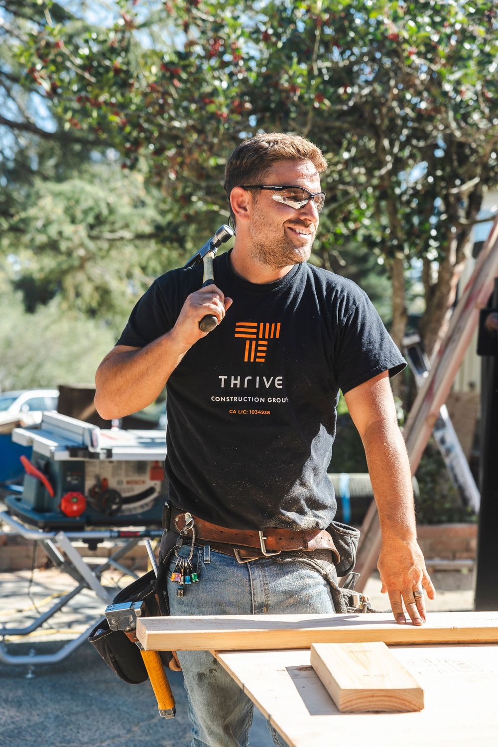 Construction worker in Thrive branded t-shirt stands with tools at outdoor worksite on sunny day.