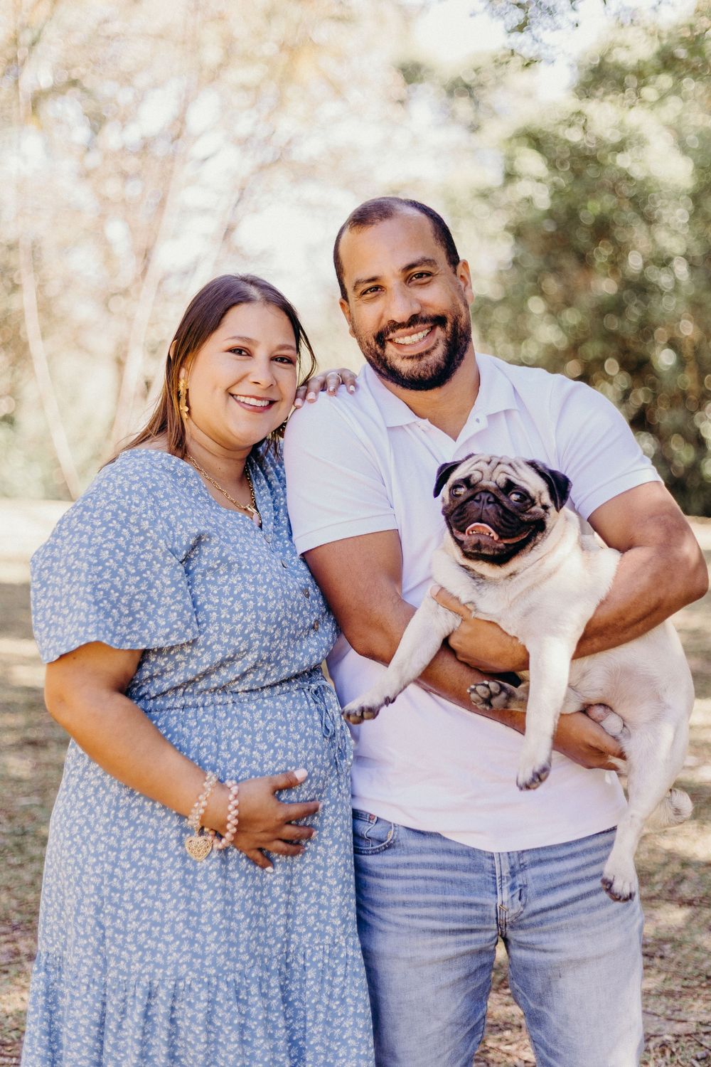 A couple in casual attire poses outdoors with their adorable pug dog on a sunny day.