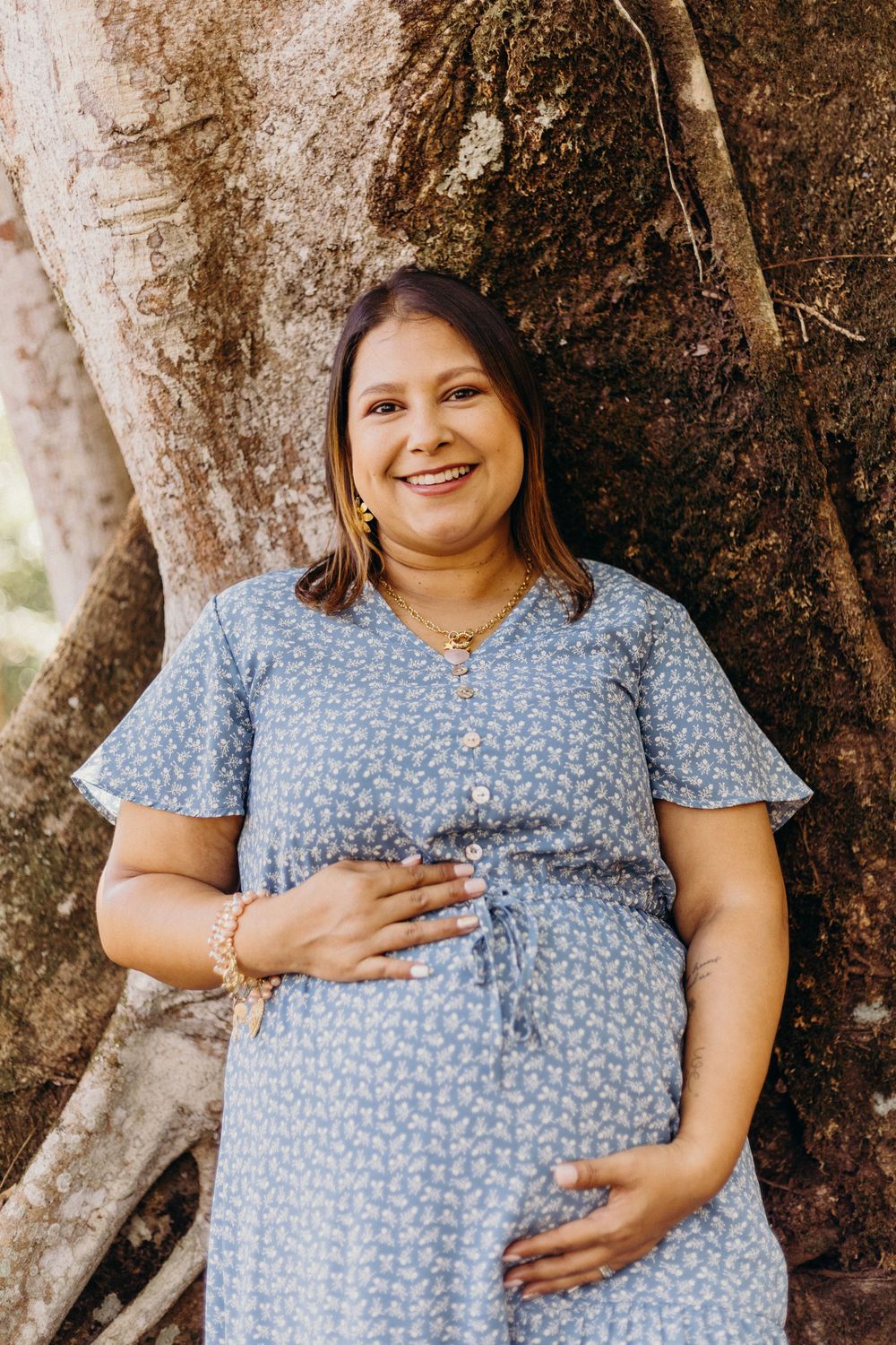 A woman in a blue floral dress poses against a tree trunk while touching her belly in maternity photos.
