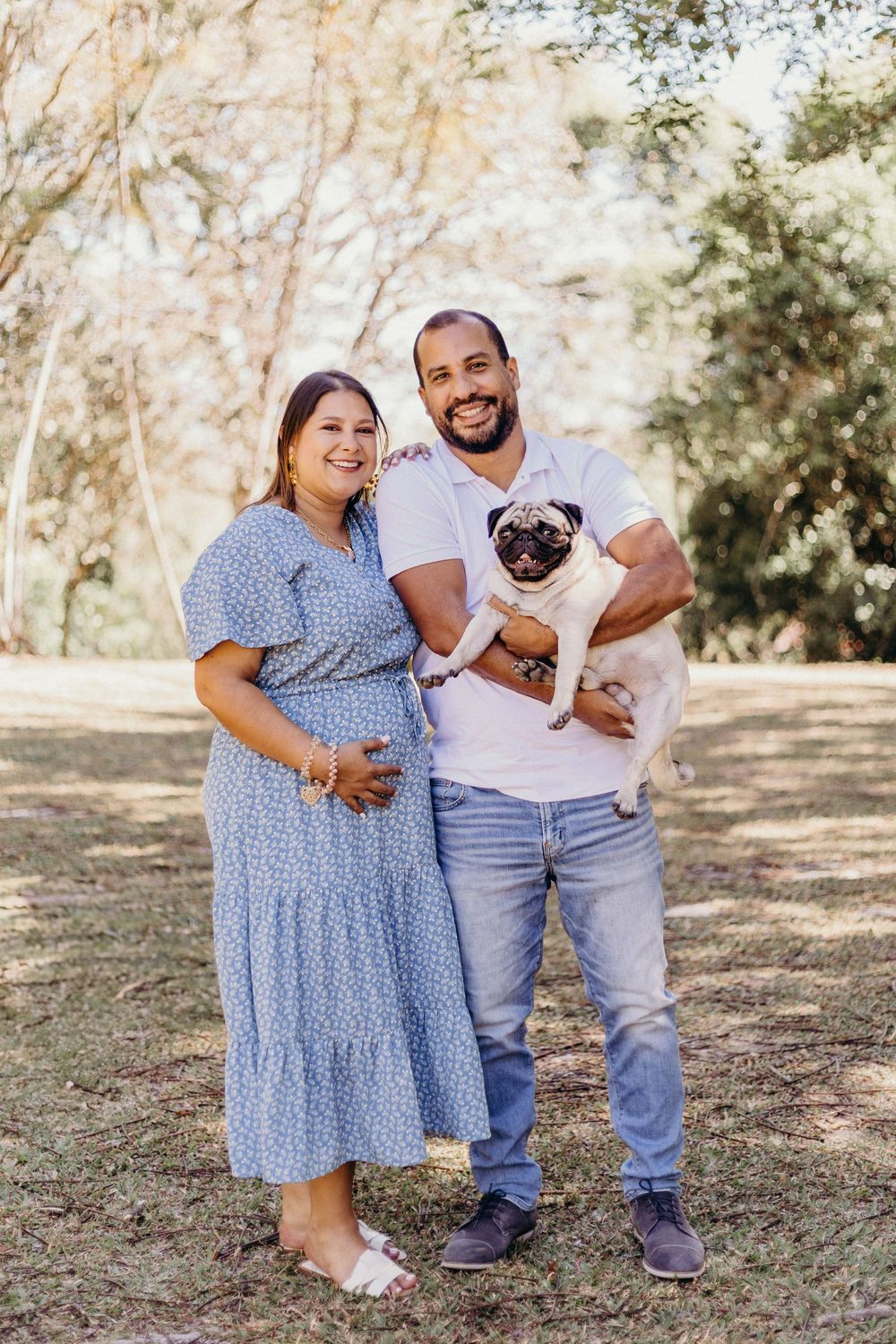 A couple sits together on grass with their pug dog while wearing coordinating blue and white outfits.