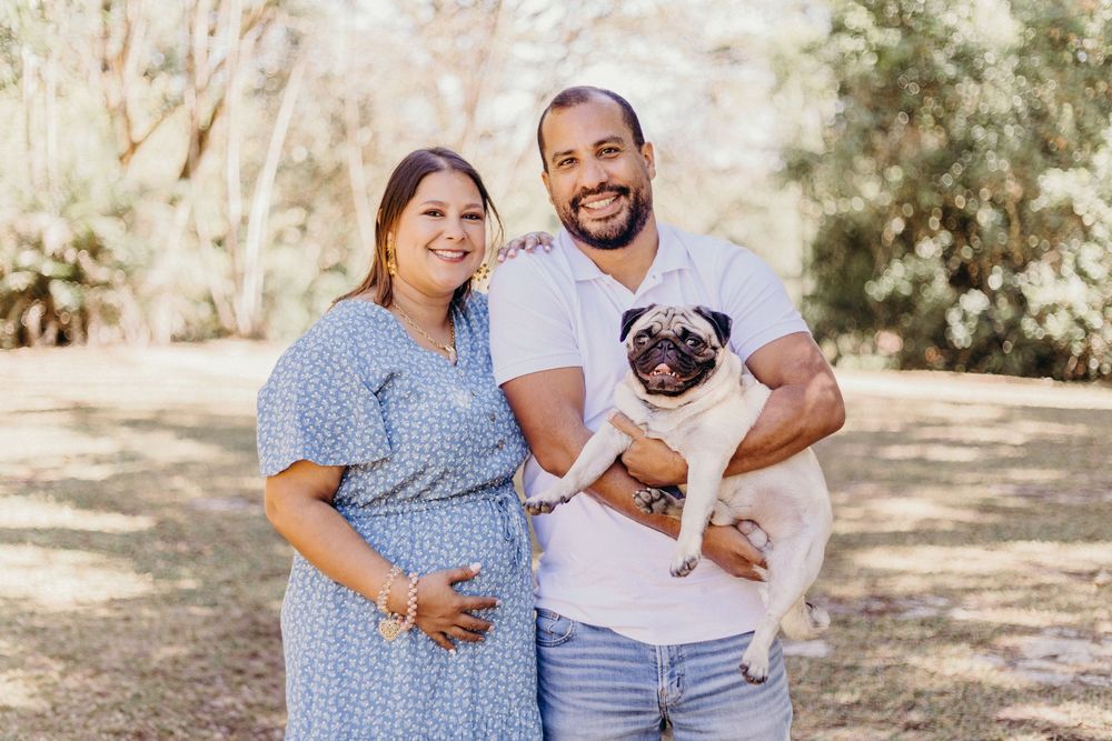 A couple poses outdoors in a sunny park setting while holding their adorable pug dog.