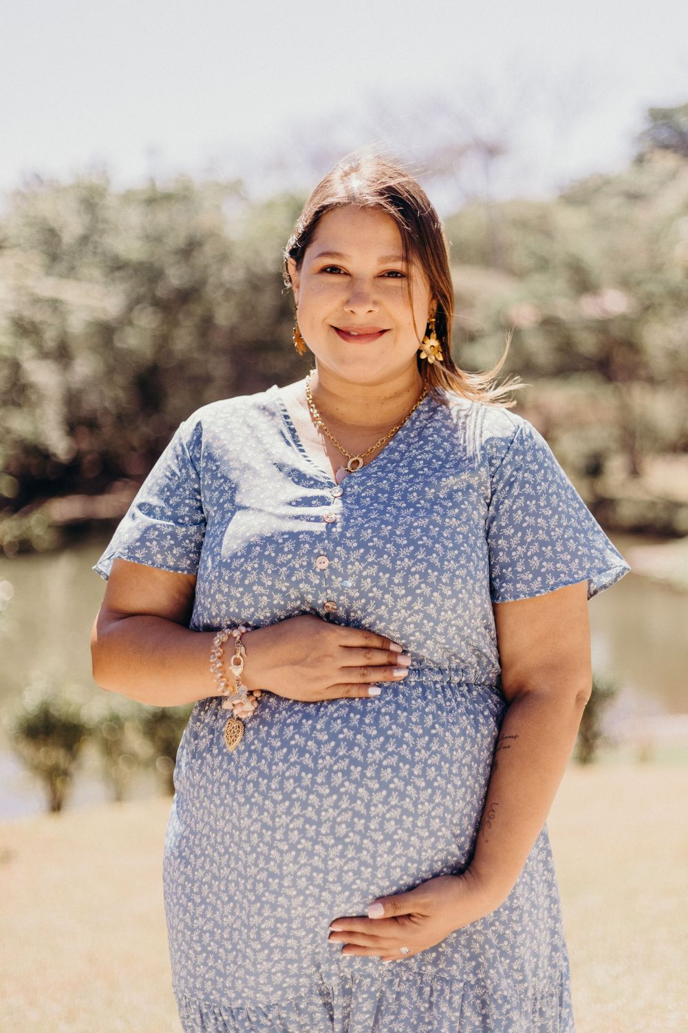 A pregnant woman in a blue floral dress smiles outdoors on a sunny day.