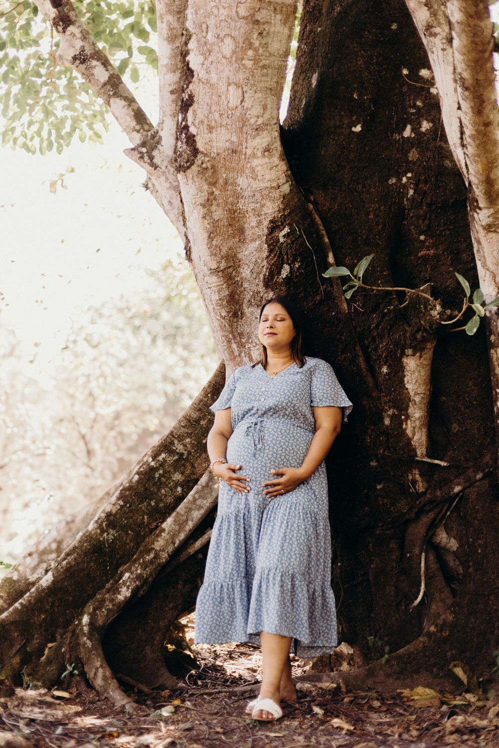 A pregnant woman in a blue polka dot dress stands against a large tree trunk in a dreamy outdoor setting.