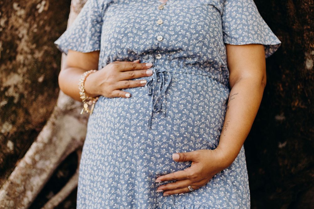 A pregnant woman in a blue floral dress cradles her baby bump while standing outdoors against a blurred natural background.