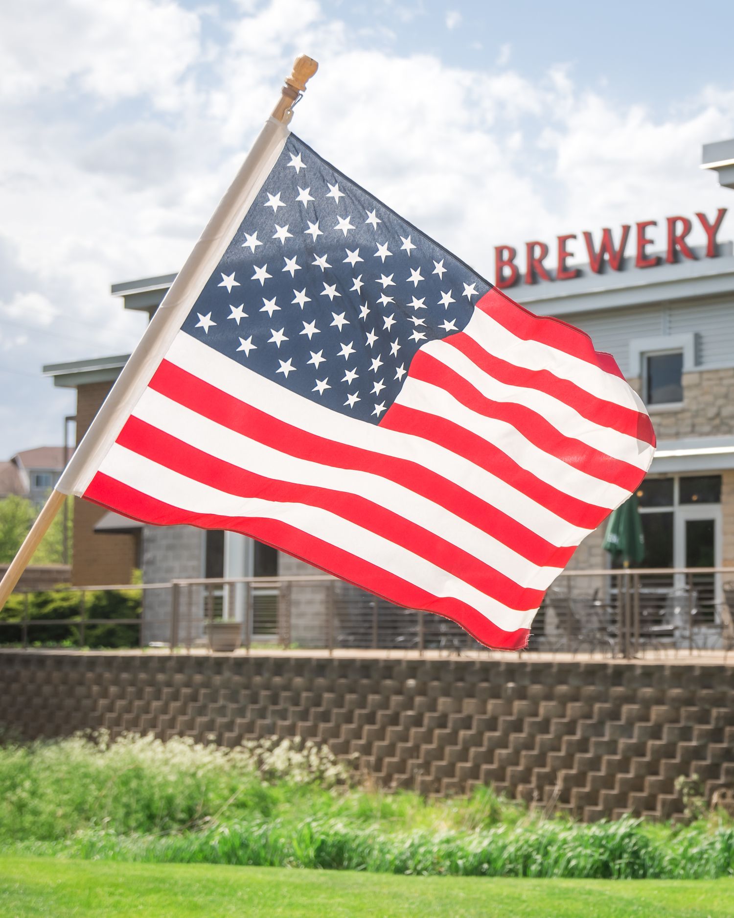 American flag waving against a cloudy sky with a brewery building visible in the background.