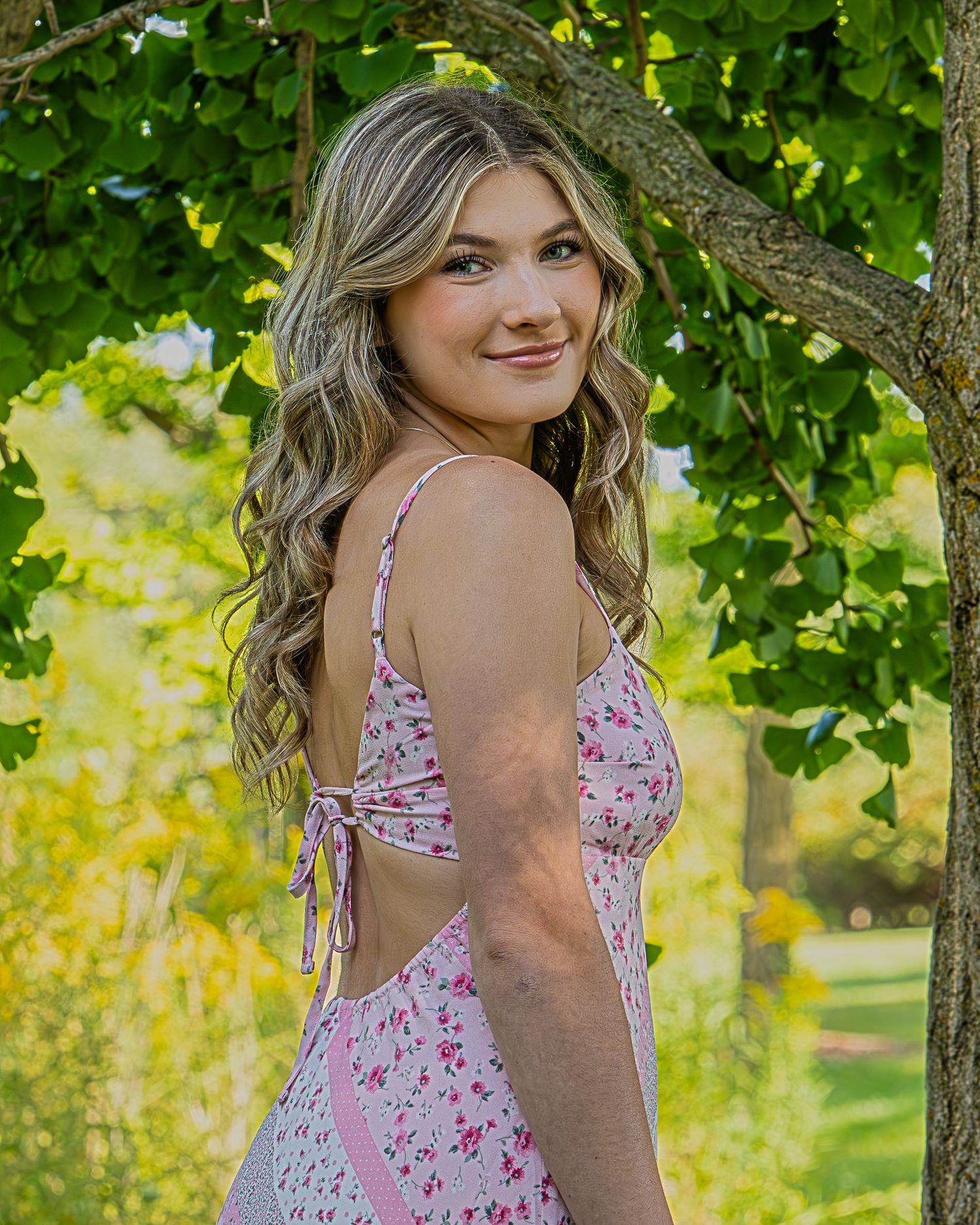 A person in a pink floral sundress poses outdoors among green foliage on a sunny summer day.