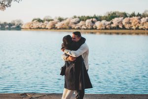 Two people embrace by the water with cherry blossom trees blooming along the shoreline during sunset.