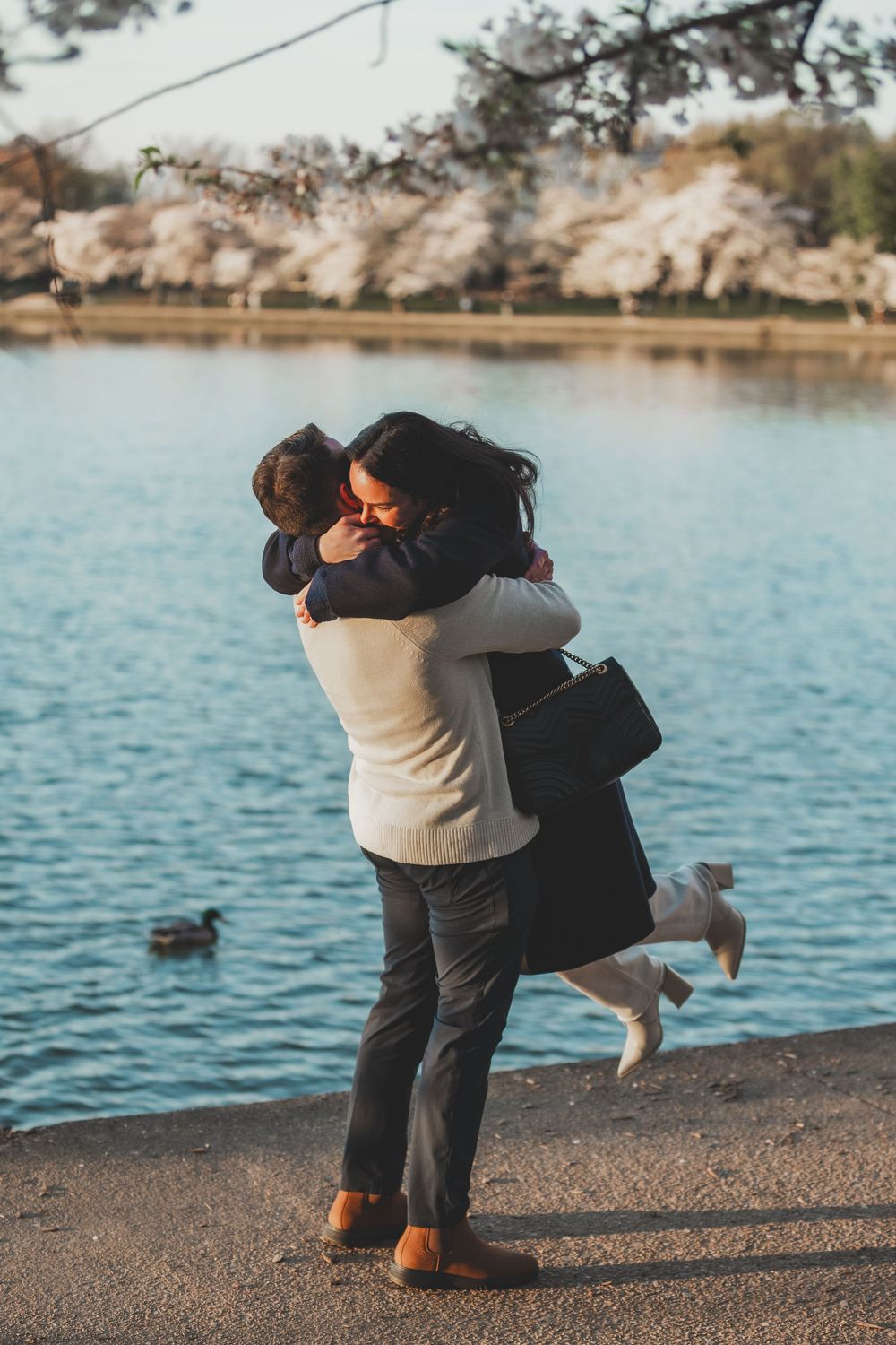 A couple embraces by a serene lake with cherry blossoms blooming in the background during springtime at the tidal basin