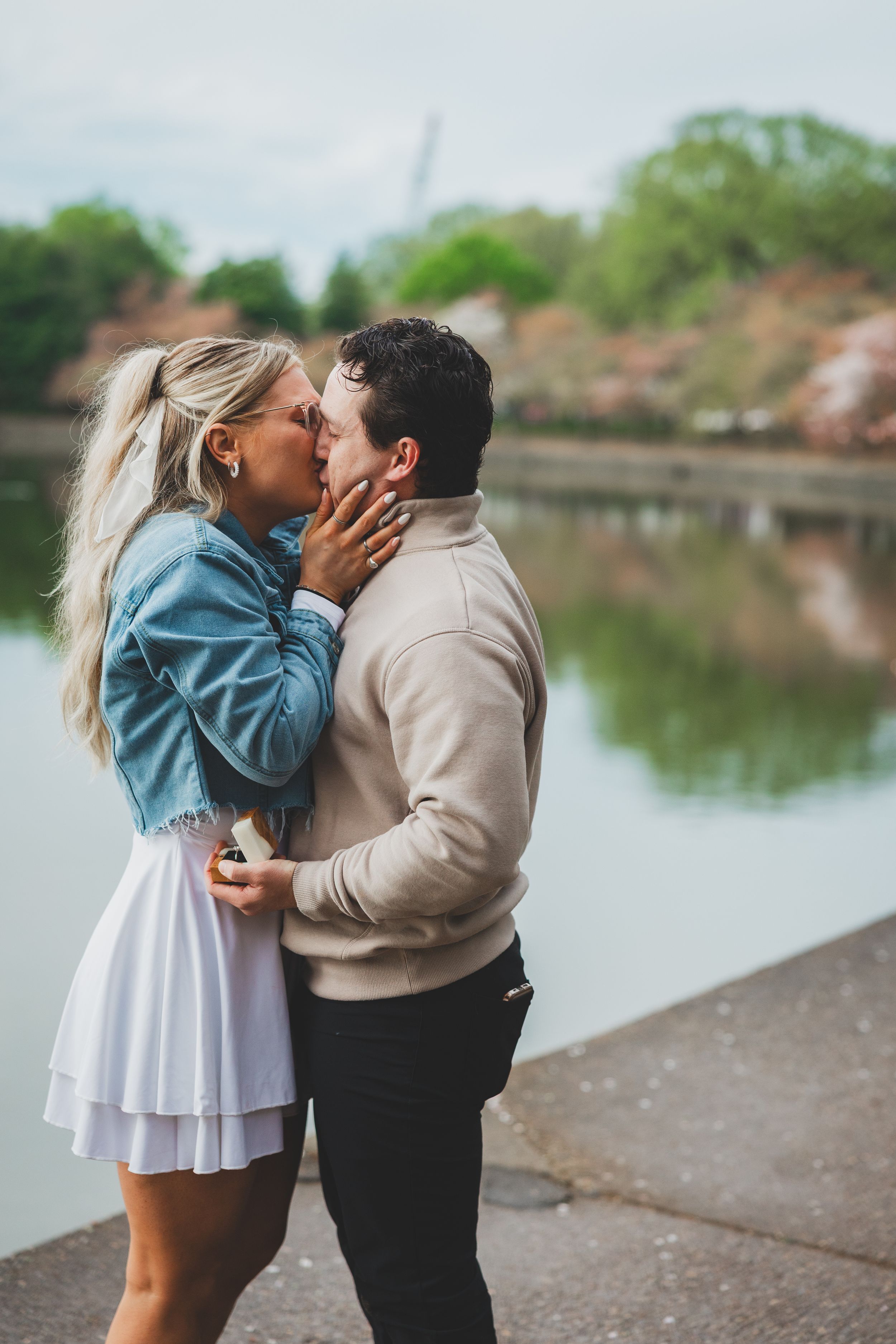 A couple shares a romantic kiss by a serene lake on a cloudy day wearing contrasting light and dark outfits.