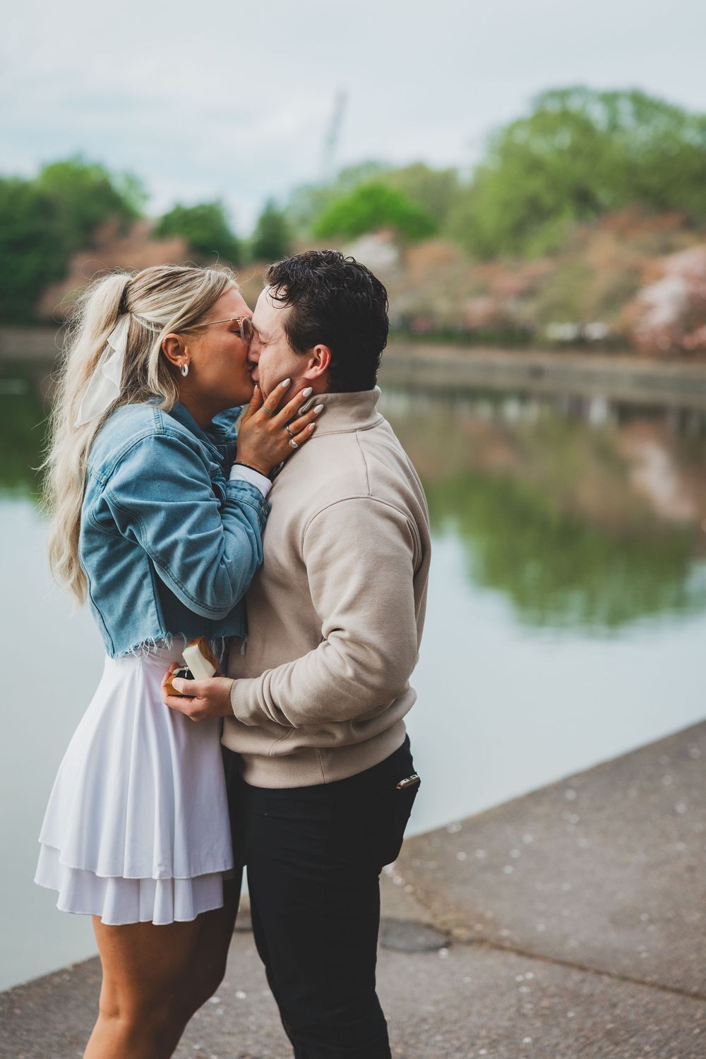 A couple shares a romantic kiss by a serene lake on a cloudy day wearing contrasting light and dark outfits.