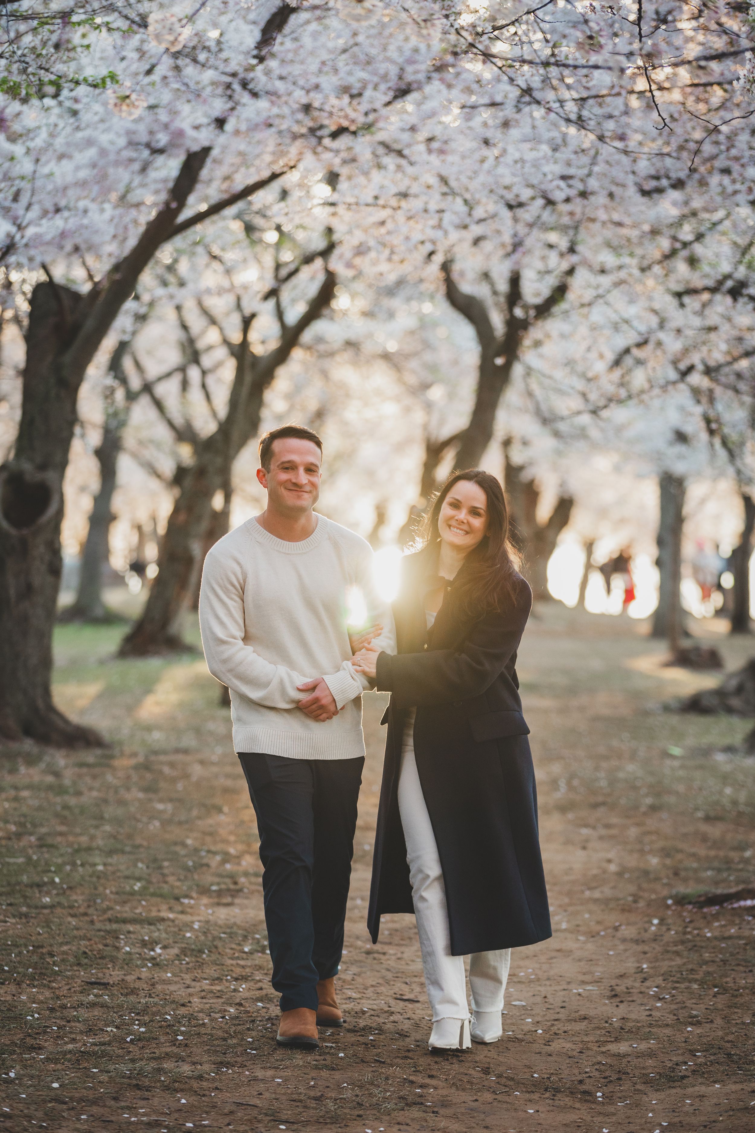 A couple laughs and walks together under blooming cherry blossom trees during sunset.