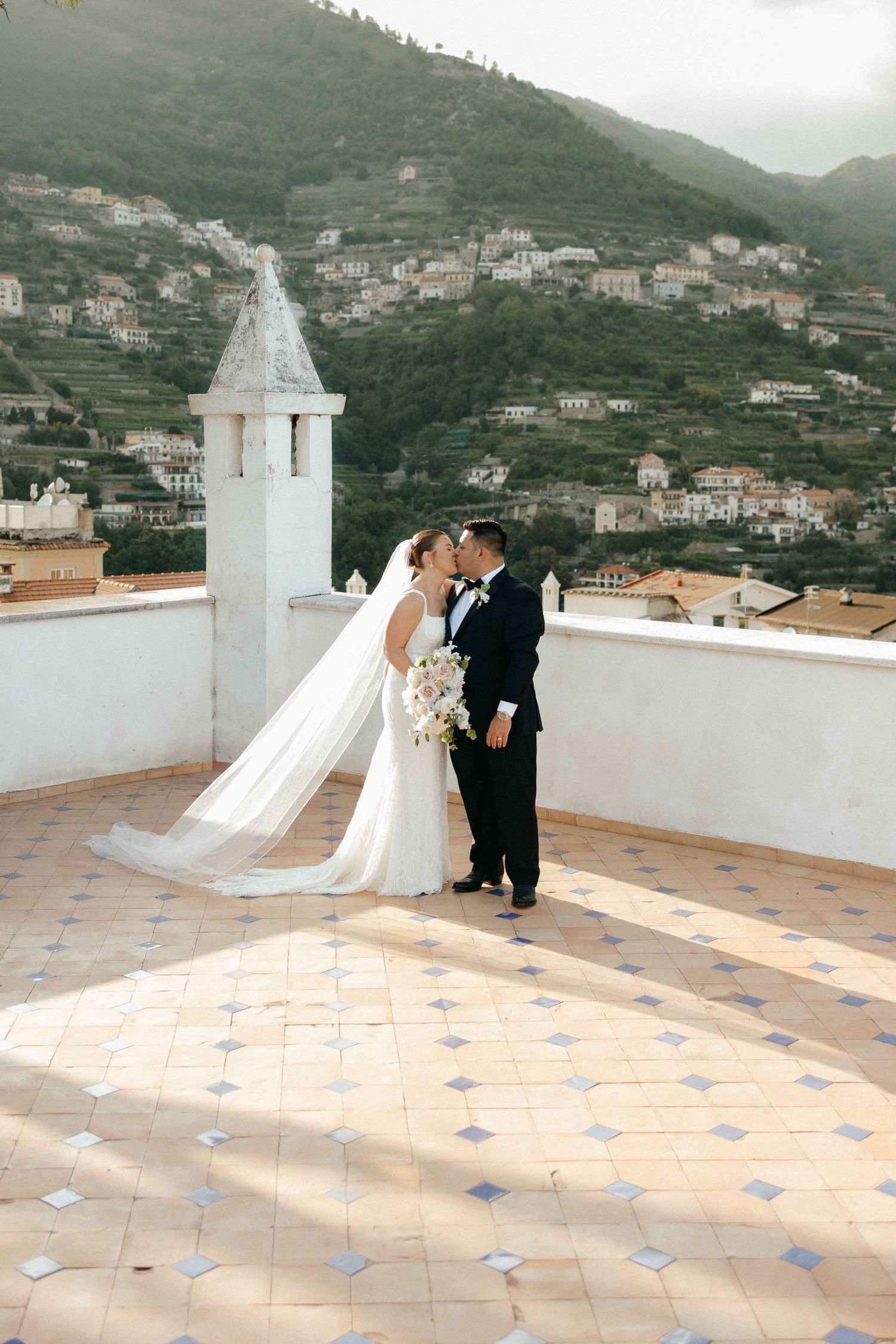 A romantic wedding couple stands on a Mediterranean terrace with tiled floors and white architecture in the background.
