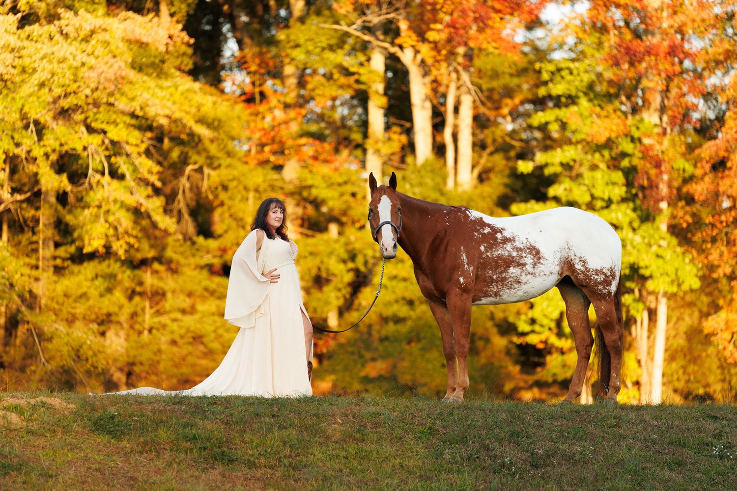 Woman in white gown standing beside chestnut pinto horse among vibrant autumn foliage.