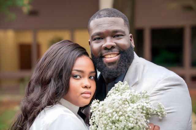 A happy couple in wedding attire poses together outdoors surrounded by greenery and white floral arrangements.