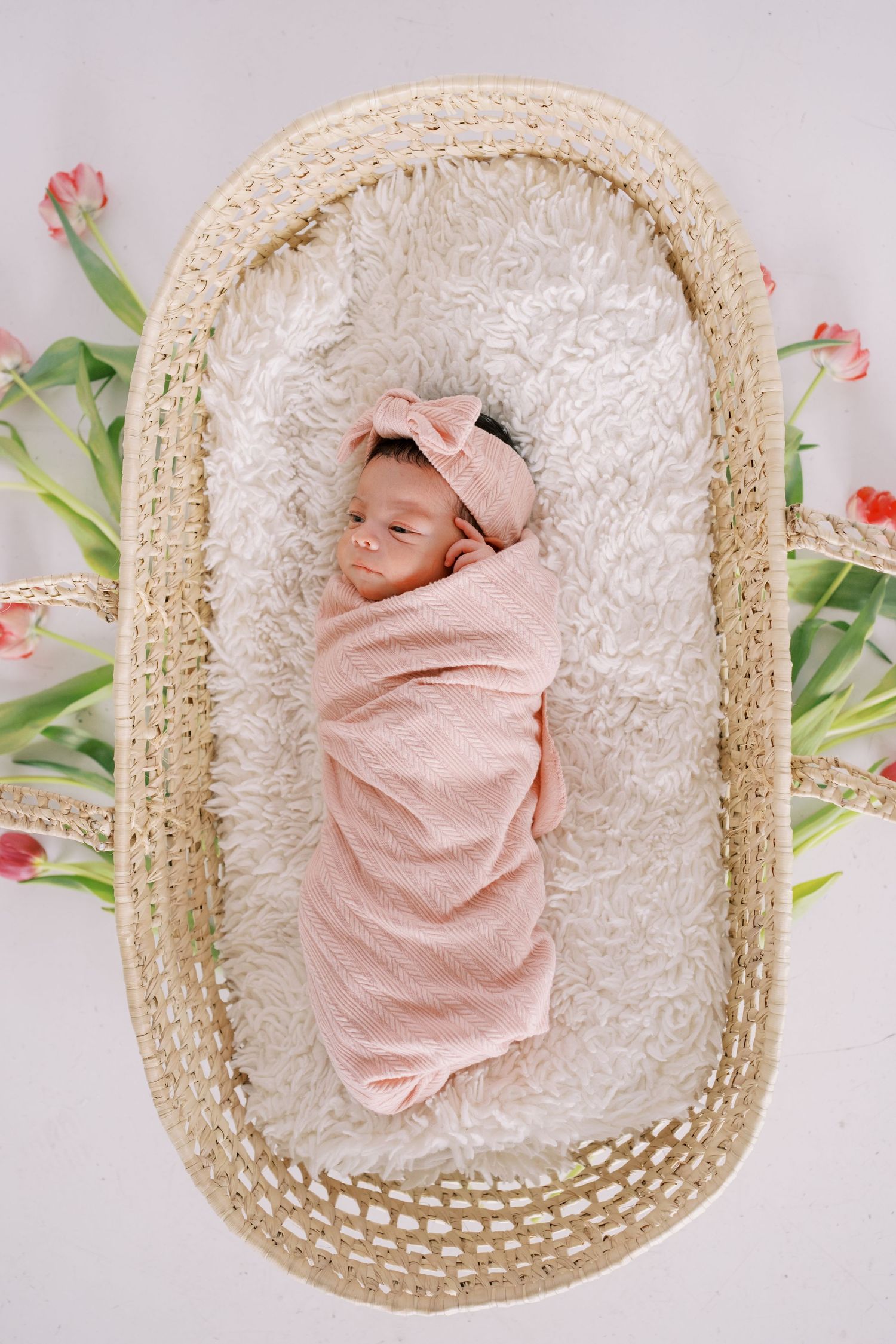 Newborn baby swaddled in pink blanket sleeping peacefully in woven basket with floral accents and white cushioning.