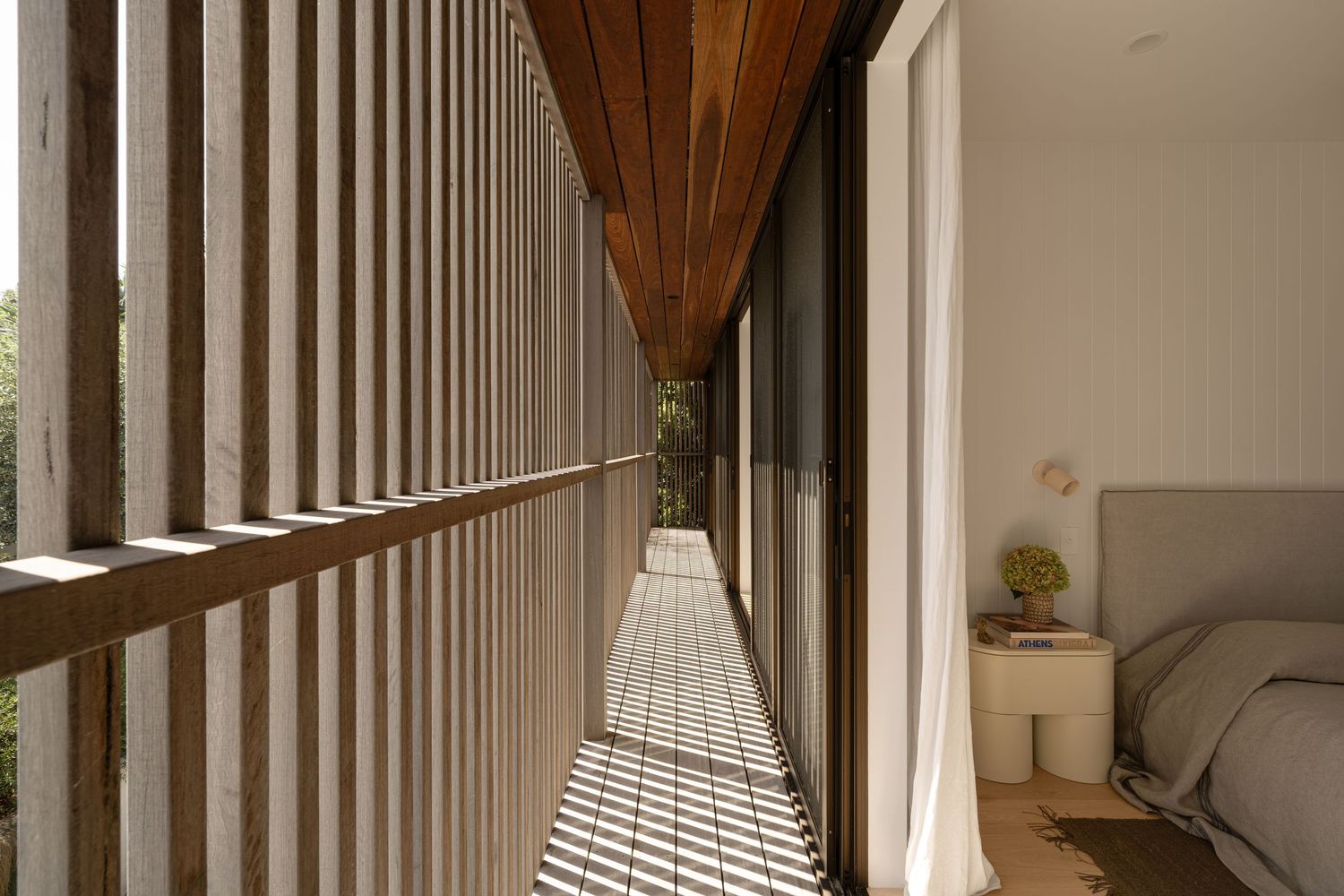 Modern bedroom interior with vertical white slats, wooden ceiling beams, and natural light streaming through windows.