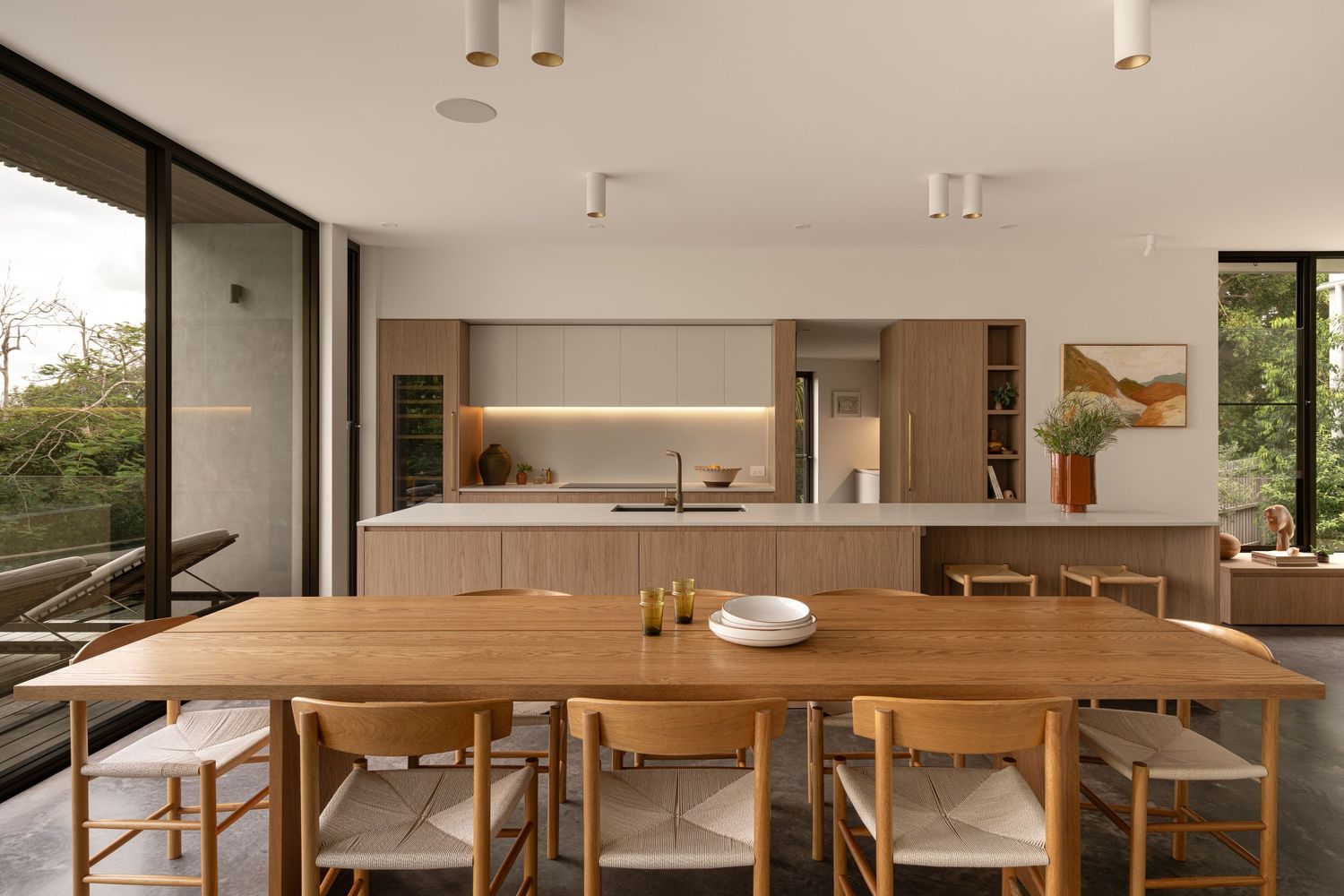 Modern dining room with wooden table and chairs connects to sleek white kitchen with floor-to-ceiling windows.