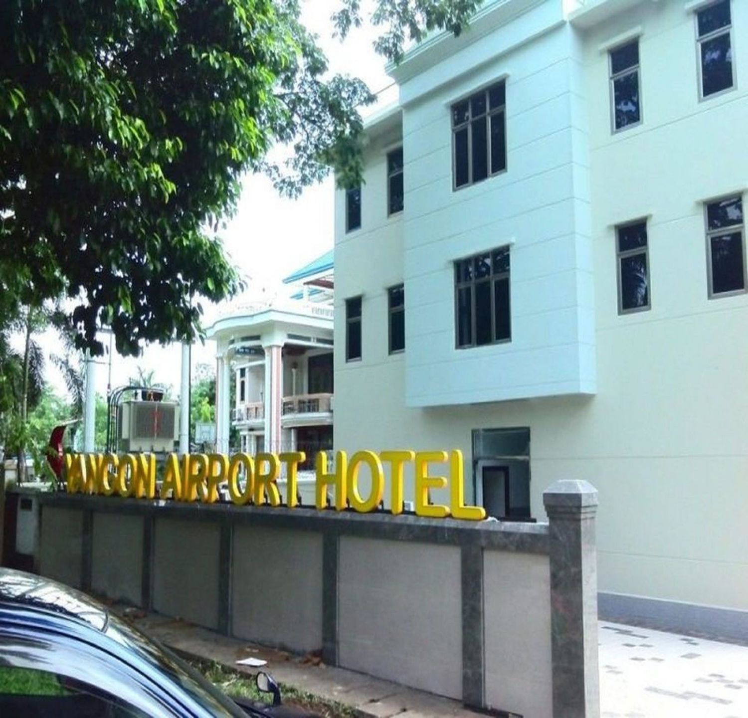 Exterior view of a white modern hotel building with tropical landscaping and parked vehicles.