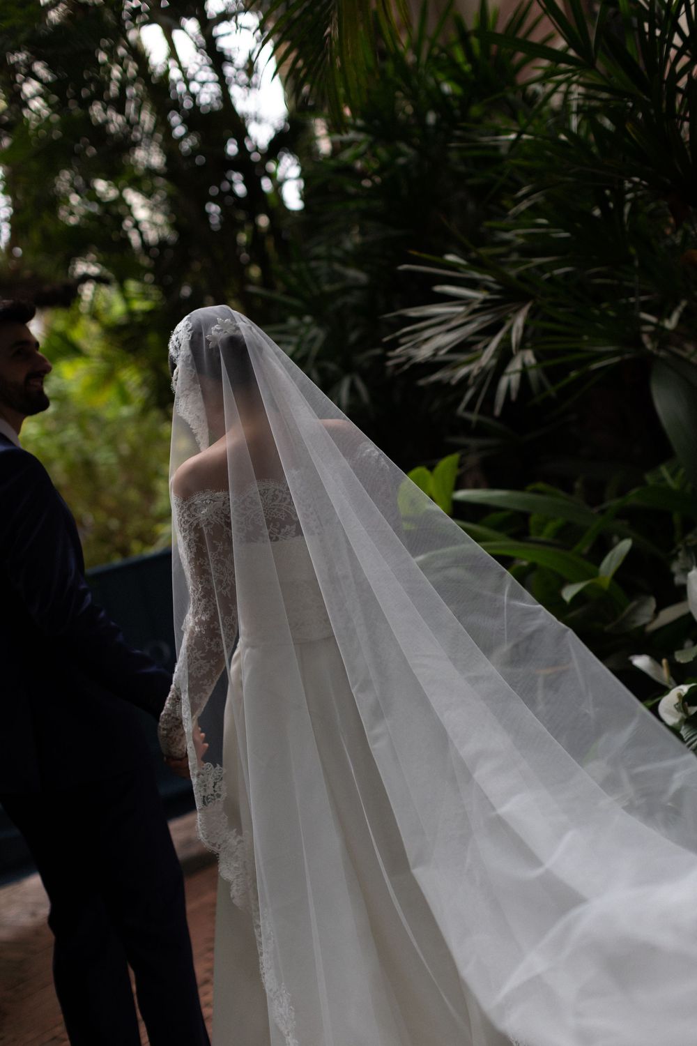 Flowing cathedral length veil trails behind white wedding dress in tropical garden setting.