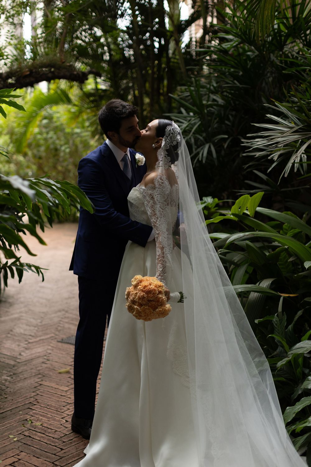 Newlyweds share an intimate moment on a brick pathway surrounded by tropical plants.