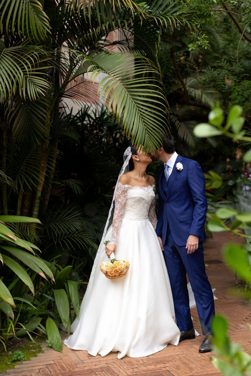 Wedding couple walking along a brick path surrounded by lush tropical palm fronds and greenery.