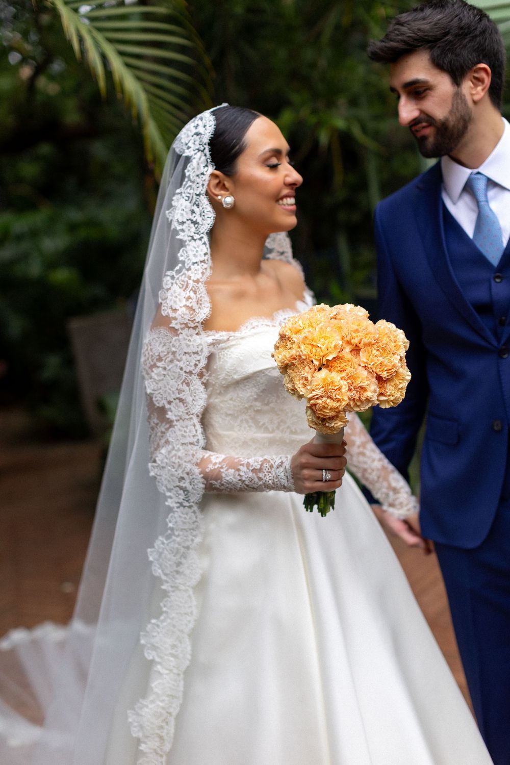Elegant bride in lace sleeve gown holds peach-colored bouquet against garden backdrop.