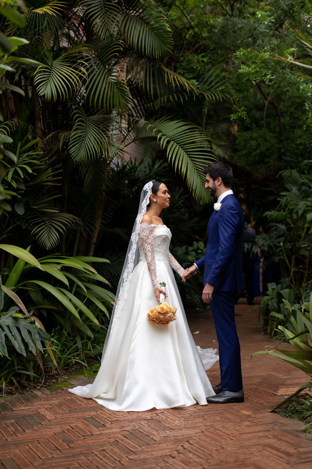 A bride and groom share a romantic moment on a brick pathway surrounded by lush tropical plants and palm fronds.