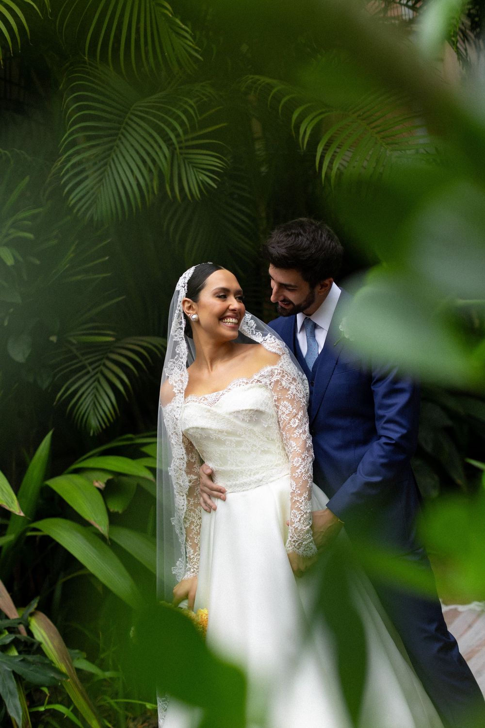 Intimate wedding portrait through tropical foliage showing couple in white dress and navy suit.