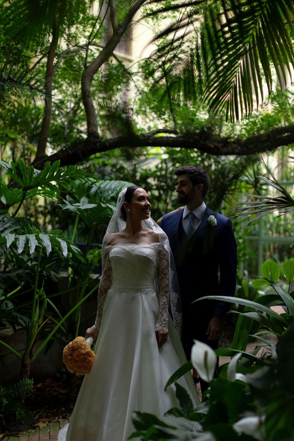 A romantic wedding couple walks through a lush tropical garden setting with palm fronds and greenery.